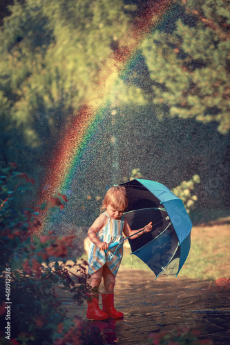 Little cute girl with blue umbrella under beautifully lighted summer rain ad bright rainbow in the park. Image with selective focus and toning