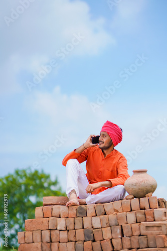 Indian farmer sitting on bricks and using smartphone at agriculture field.