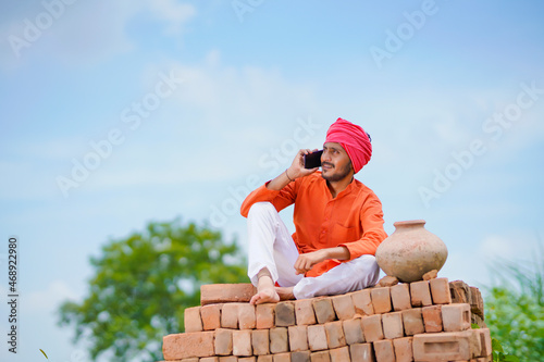 Indian farmer sitting on bricks and talking on smartphone at green corn field.