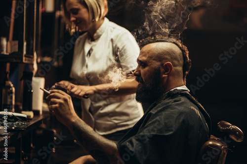 Young bearded man with a mohawk getting haircut by hairdresser and smoking a cigarette