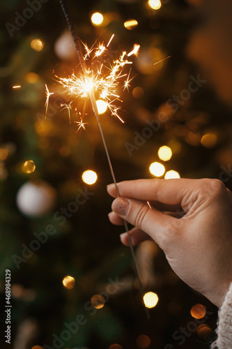 Hand holding burning sparkler against christmas tree lights in evening room. Happy New Year!