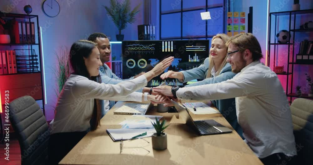 Team trust support of attractive smiling confident experienced diverse male and female office workmates which sitting behind the table and making unity symbol with their hands in evening office