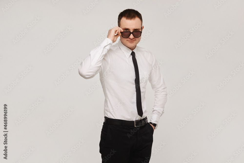 A young successful confident guy in a white shirt and a black tie adjusts his sunglasses on a gray background. Isolated with copy space A young successful confident guy in a white shirt and a black tie adjusts his sunglasses on a gray background. Isolated with copy space