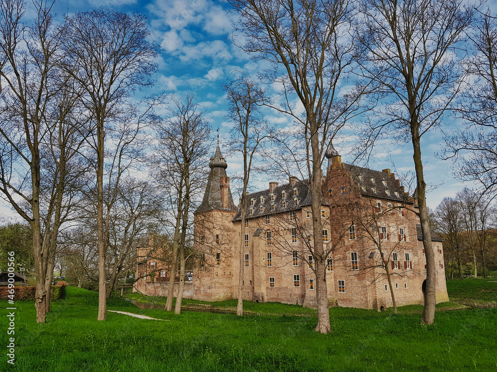 Fototapeta premium The medieval castle of Doorwerth on the river Rhine in the Netherlands, with green grass and large trees in the foreground. The castle was severely damaged in WW2, but has been restored 