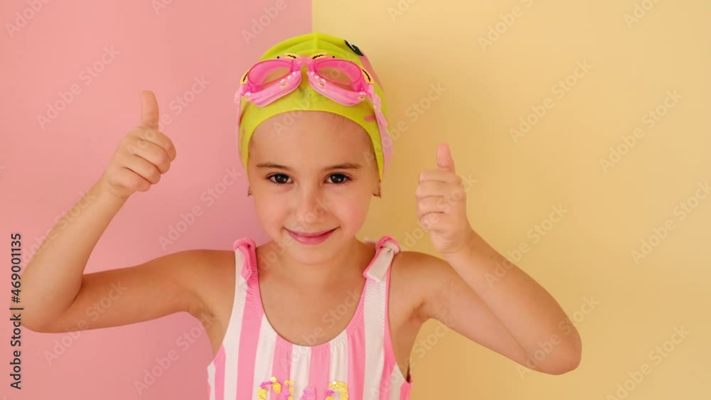 Child swimmer showing success gesture, thumb up with hands, saying well done. Baby girl in swimming goggles, a yellow swimming cap and a striped swimsuit with rhinestones. 