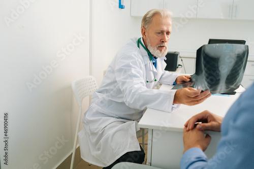 View from back of unrecognizable male patient listening mature adult doctor with chest x-ray lungs scan giving professional medical consultation for treatment illness  explaining causes of disease.