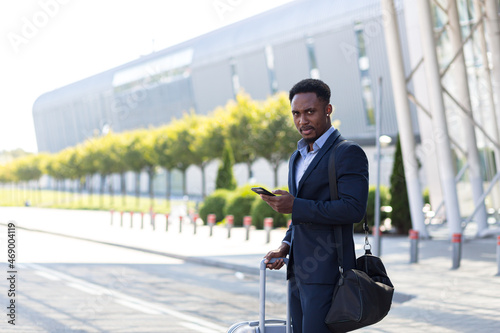 african american business man standing on the background a modern train station airport in formal suit with a suitcase using app mobile phone. Traveling businessman tourist with a smartphone in hands