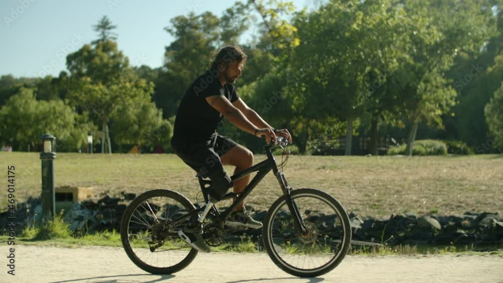 Side view of young Caucasian man with disability cycling in park. Tracking shot of adult with bionic leg riding bicycle, taking care of health by doing physical activity. Disability, sport concept Side view of young Caucasian man with disability cycling in park. Tracking shot of adult with bionic leg riding bicycle, taking care of health by doing physical activity. Disability, sport concept