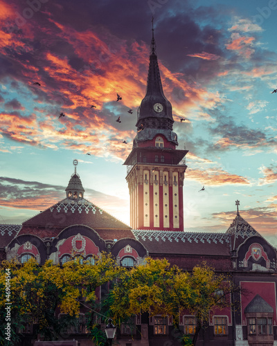 Pigeons flying past the magnificent tower of Subotica's city hall with fiery sky during sunset