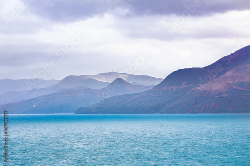 Turquoise water surrounding a small coral reef close to the shore with mountains in the background covered in haze and clouds.