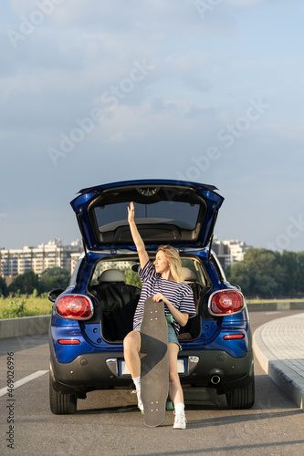 Happy woman travel by car relax sitting in trunk with longboard enjoy sunset city view during summer vacation. Carefree female love freedom and relaxation on road trip. Traveler lifestyle concept © DimaBerlin