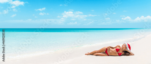 Christmas luxury vacation woman lying down on perfect beach sunbathing in sun during winter travel xmas holidays. Bikini model wearing santa hat for wellness promotion, body skincare, spa treatment.