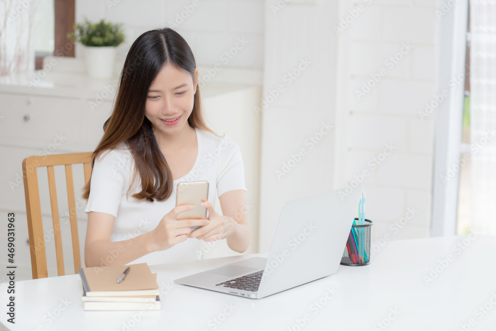 Young asian woman working laptop computer and reading smartphone on internet online on desk at home, freelance girl smiling using phone with social media, business and communication concept. Young asian woman working laptop computer and reading smartphone on internet online on desk at home, freelance girl smiling using phone with social media, business and communication concept.