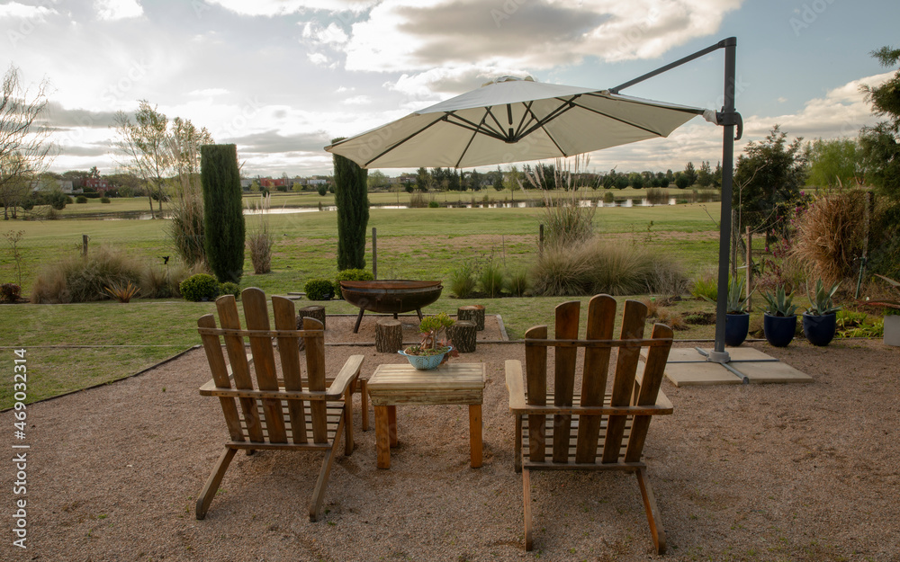 Relaxation. View of the beautiful garden with wooden exterior furniture, garden chairs and table, and big sunshade umbrella at sunset.