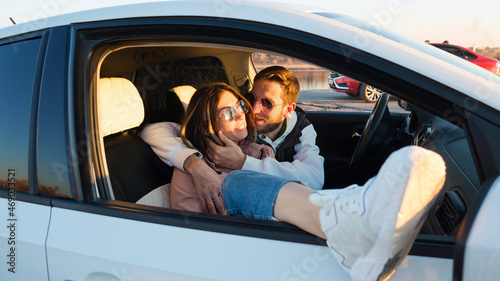 Loving carefree tourists travelers couple sitting in car at sunset in countryside. Man hugs and kisses his girlfriend while stopping on trip. Travel, road trip and comfortable concept © Vagengeim