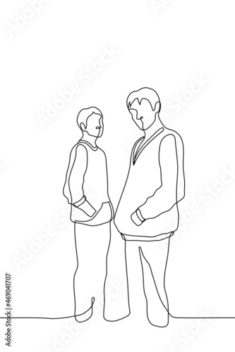 two men in hoodies are standing sideways with their hands in their pockets and looking at the viewer - one line drawing vector. concept of two male friends; popularity of hoodies among young people