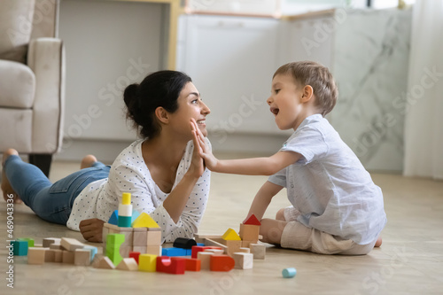 Happy nanny giving high five praise to excited preschool kid boy for completing toy tower on warm floor. Indian babysitter and kid playing at home, constructing building from small wooden blocks photo