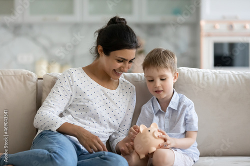Happy mom teaching little son to save money, keeping reserve fund. Mother and kid dropping cash into pink piggy bank, playing financial game on sofa, counting budget, planning child future photo