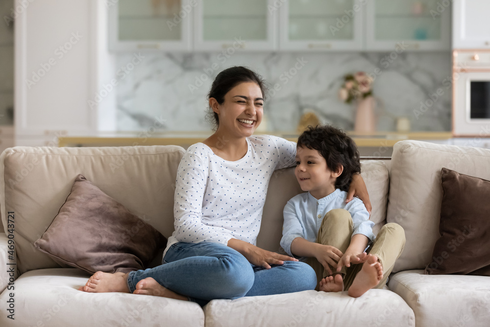 Laughing Indian mom enjoying leisure time with happy son at home, talking to boy. Mother and cheerful preschool child having fun together, relaxing on couch. Babysitter watching cute kid