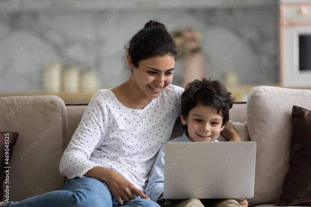 Happy Indian little son and loving mom enjoying leisure at laptop on couch watching movie using