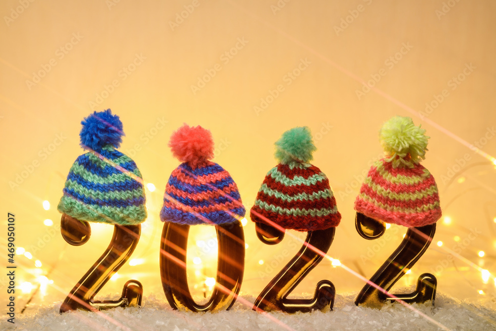Gold numbers 2022 with multi-colored knitted hats with anamorphic filter and luminous garland on yellow background. New year backdrop with 2022 year