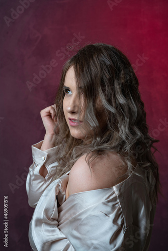 Super sexy, happy, fashionable and stylish girl plus size poses in a satin blouse with a deep neckline and bare shoulder, gray trousers and jewelry accessories on a red-pink gradient background 