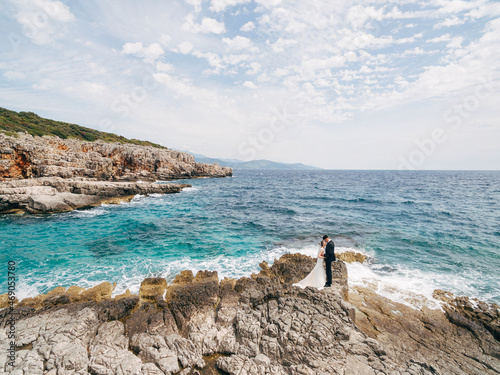 Bride and groom stand embracing on the rocky shore against the backdrop of the seething sea