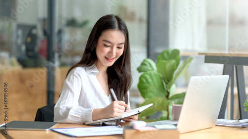 Pretty young Asian businesswoman working on laptop and taking notes In office..