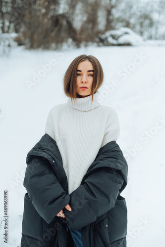 Attractive girl in a sweater stands on a snowy street in the cold and poses for the camera with a serious face with his jacket down.