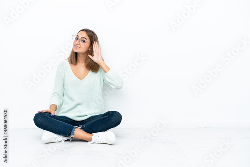 Young caucasian woman sitting on the floor isolated on white background listening to something by putting hand on the ear