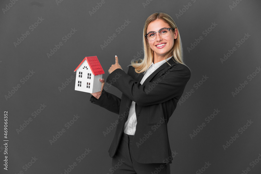 Blonde Uruguayan girl holding a house toy isolated on black background pointing back Blonde Uruguayan girl holding a house toy isolated on black background pointing back