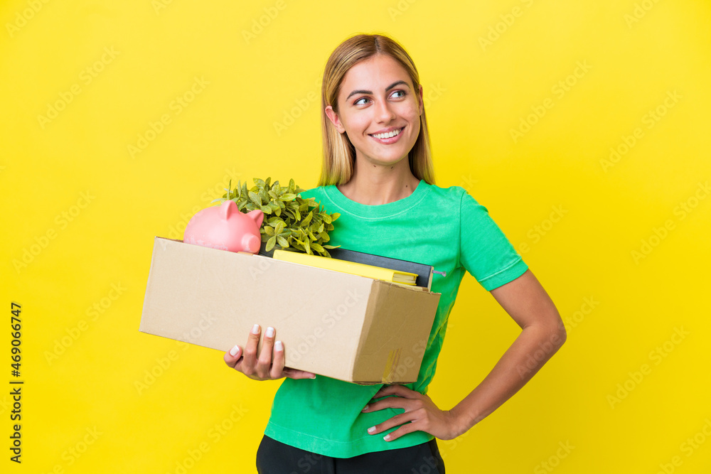 Young Uruguayan girl making a move while picking up a box full of things isolated on yellow background posing with arms at hip and smiling Young Uruguayan girl making a move while picking up a box full of things isolated on yellow background posing with arms at hip and smiling