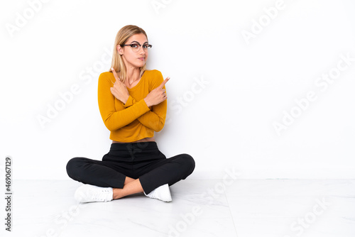 Blonde Uruguayan girl sitting on the floor pointing to the laterals having doubts