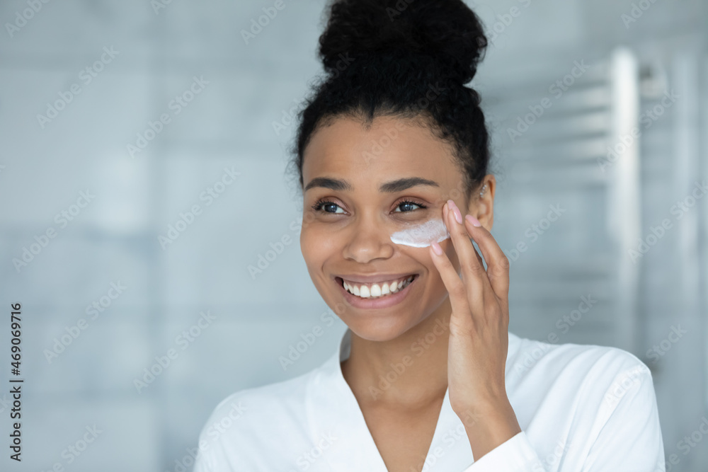 Smiling attractive millennial african american biracial woman in bathrobe applying moisturizing creme, enjoying taking care of facial skin, doing daily beauty routine at home., product ad concept. Smiling attractive millennial african american biracial woman in bathrobe applying moisturizing creme, enjoying taking care of facial skin, doing daily beauty routine at home., product ad concept.
