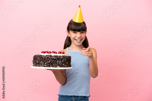Little caucasian kid holding birthday cake isolated in pink background surprised and pointing front