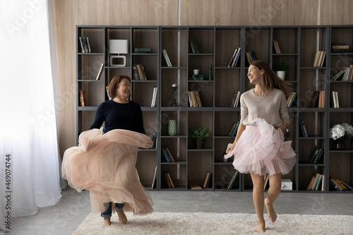 Happy woman with mature mother wearing pink princess skirts dancing having fun at home together, smiling senior mom with grown-up daughter moving to favorite music, family engaged in funny activity photo