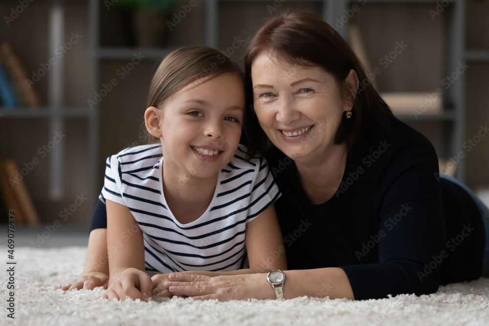 Head shot portrait of smiling mature grandmother and little granddaughter hugging lying on warm floor at home, happy senior woman with adorable girl kid grandchild looking at camera, two generations