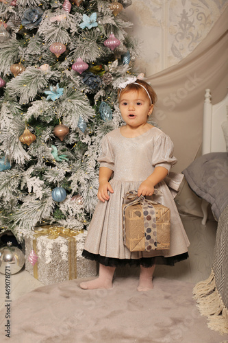 a beautiful little girl in a silver dress stands at the Christmas tree at home and holds a gift box in her hands