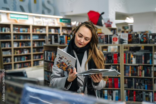 A beautiful young woman chooses a comic book for a gift in a bookstore to buy it. Stands in front of a bookcase and holds two comics comparing them.