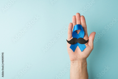 First person top view photo of male hand holding blue ribbon and mustache shape in palm symbol of prostate cancer awareness on isolated pastel blue background with empty space photo