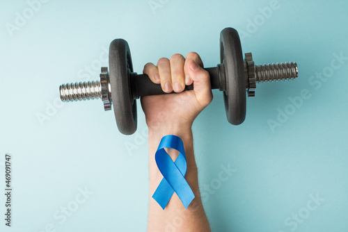 First person top view photo of raised man's hand holding dumbbell and blue ribbon on wrist symbol of prostate cancer awareness on isolated pastel blue background