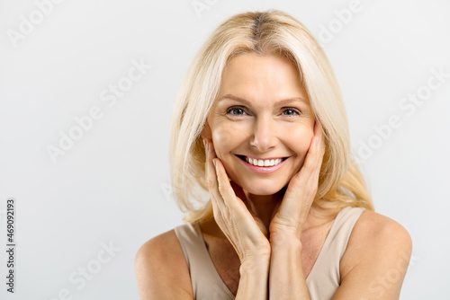 Enchanting middle-aged woman with blonde hair hug herself and look at the camera over grey background, mature lady smiles widely and gently touch face with clear skin, fine wrinkles near eyes