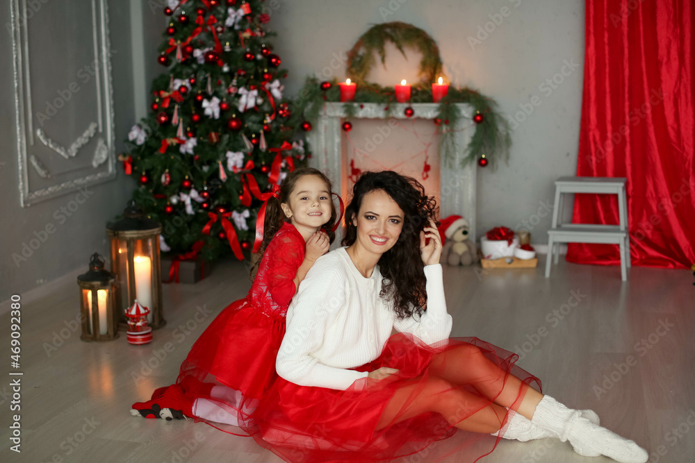 Happy mom celebrates Christmas with her daughter. Mom and daughter are dressed in red skirts and the Christmas tree is decorated in multi-colored lights. Happy mom celebrates Christmas with her daughter. Mom and daughter are dressed in red skirts and the Christmas tree is decorated in multi-colored lights.