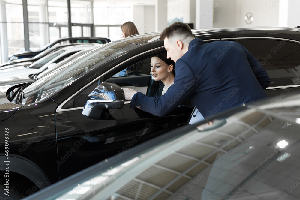 Side view of young beautiful woman sitting inside car and holding hand on steering wheel. She smiling and talking with manager of car dealership. Car agent representing inside of automobile. Side view of young beautiful woman sitting inside car and holding hand on steering wheel. She smiling and talking with manager of car dealership. Car agent representing inside of automobile.