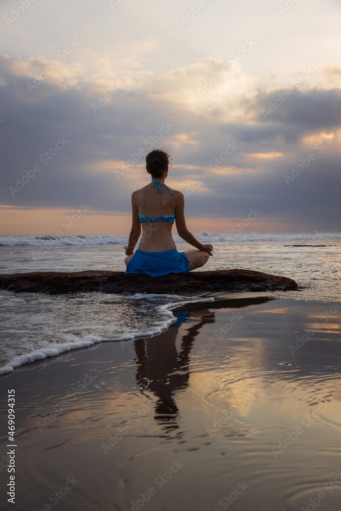 Sunset yoga. Caucasian woman sitting on the stone in Lotus pose. Padmasana. Hands in gyan mudra. Beach in Bali. View from back. Copy space. Yoga retreat. Water reflection. Self care. Zen life.