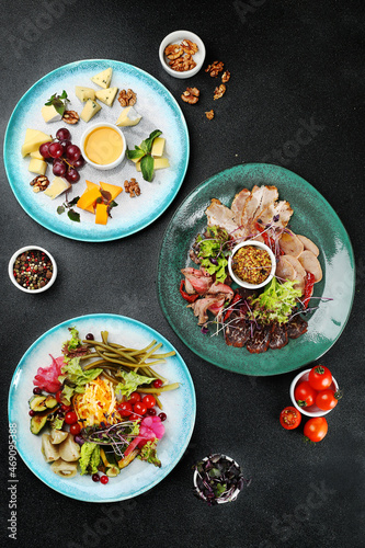 charcuterie table with three plate on dark concrete background top view. cheese plate with honey and grape, asorted pickled vegetables plate antipasti, ham plate