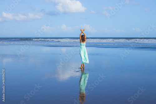 Young woman walking barefoot on empty beach. Full body portrait. Slim Caucasian woman wearing long dress. View from back. Water reflection. Hands raised up. Vacation in Asia. Travel concept.