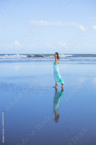 Young woman walking barefoot on empty beach. Full body portrait. Slim Caucasian woman wearing long dress. Water reflection. Summer sun light. Vacation in Asia. Travel concept. Bali, Indonesia