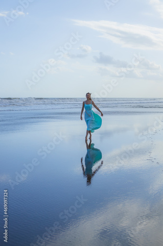 Happy smiling woman walking barefoot on empty beach. Full body portrait. Slim Caucasian woman wearing long dress. Water reflection. Blue sky. Vacation in Asia. Travel concept. Bali  Indonesia