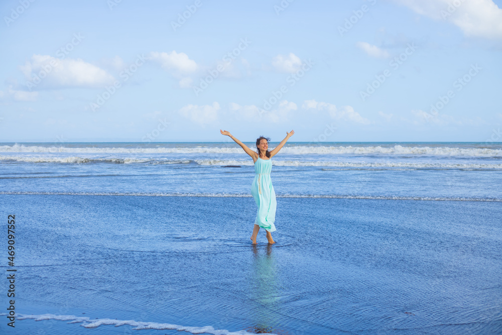 Young woman walking barefoot on empty beach. Full body portrait. Slim Caucasian woman wearing long dress. Hands raised up. Water reflection. Freedom concept. Summer vacation in Asia. Bali Young woman walking barefoot on empty beach. Full body portrait. Slim Caucasian woman wearing long dress. Hands raised up. Water reflection. Freedom concept. Summer vacation in Asia. Bali